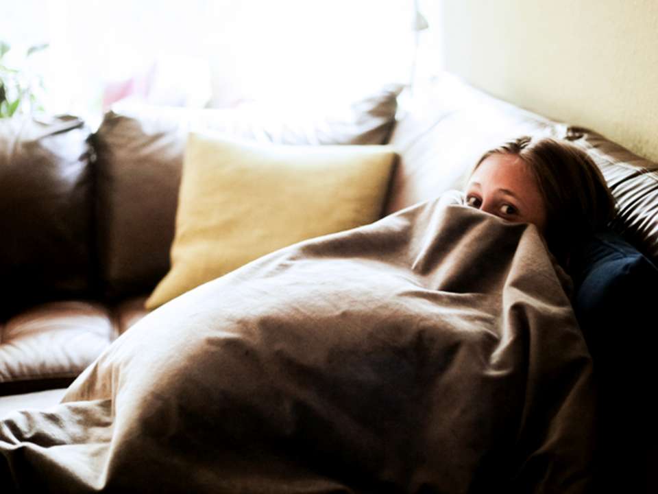 A woman sits on a couch hiding under a blanket.