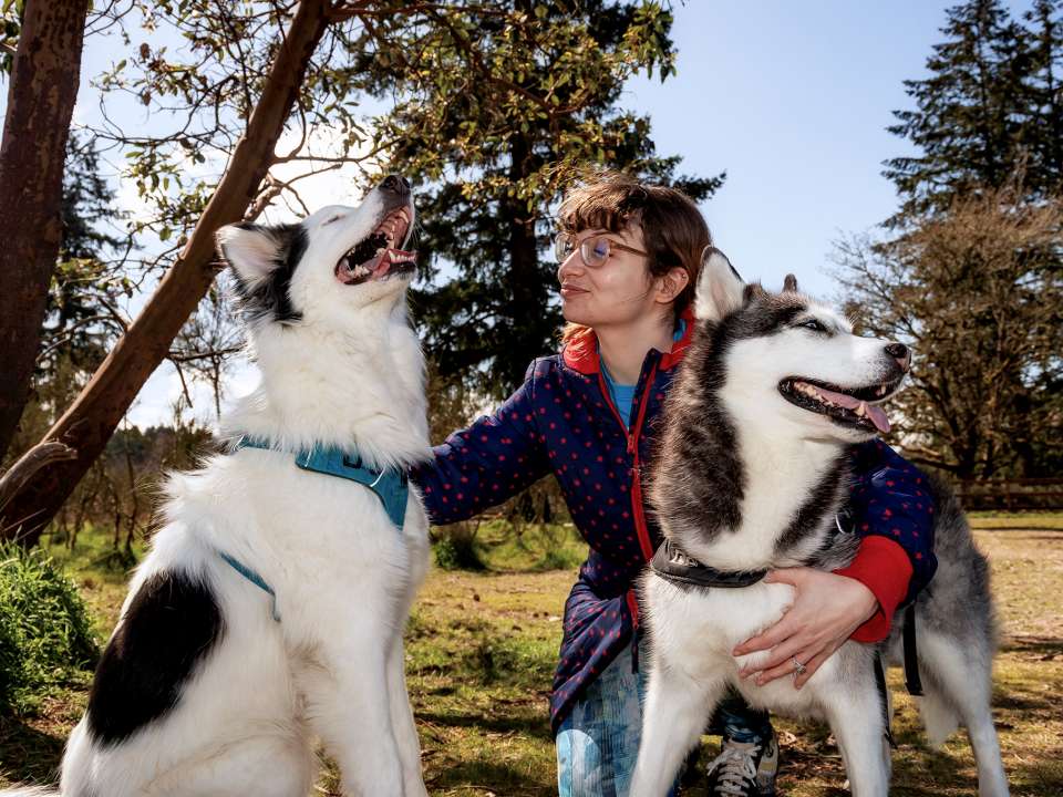A woman kneeling outdoors with two husky-like dogs.