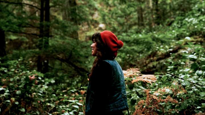 A woman with a red hat stands in a green forest.