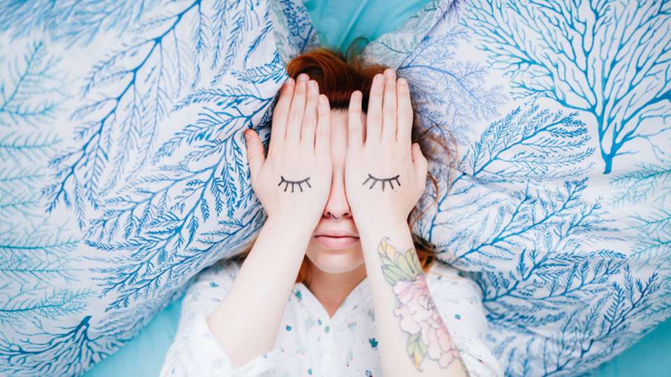 woman lying in bed covering her eyes with her hands with eyebrows painted