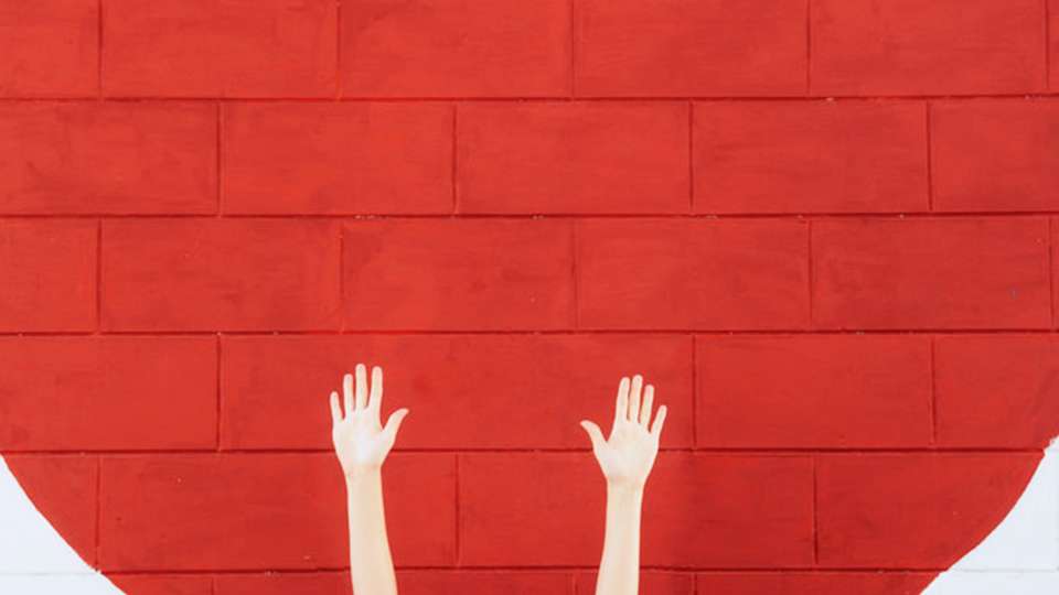 Hands reaching up in front of a brick wall painted red.