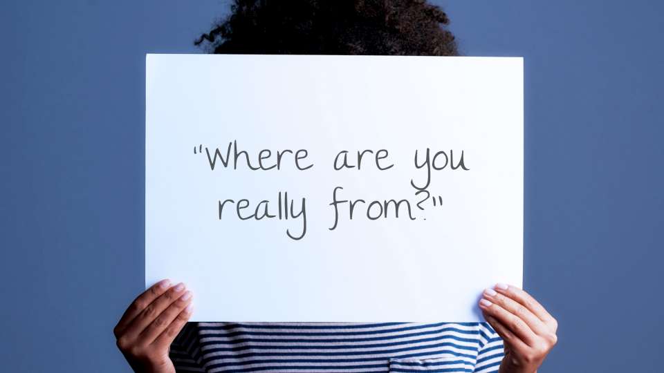 A black woman holding a sign with a microaggression written on it that says: Where are you really from?.