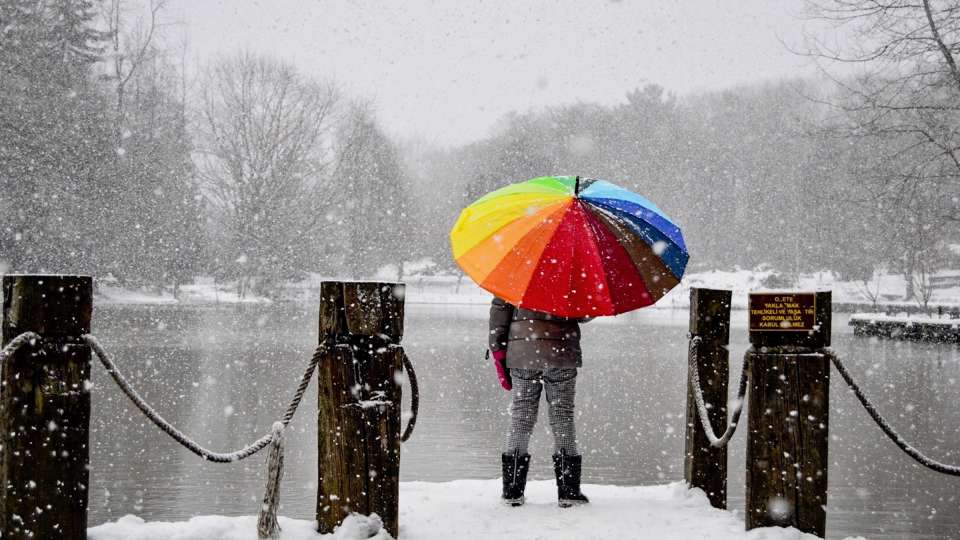 Person standing in snow by a lake, holding a rainbow umbrella.