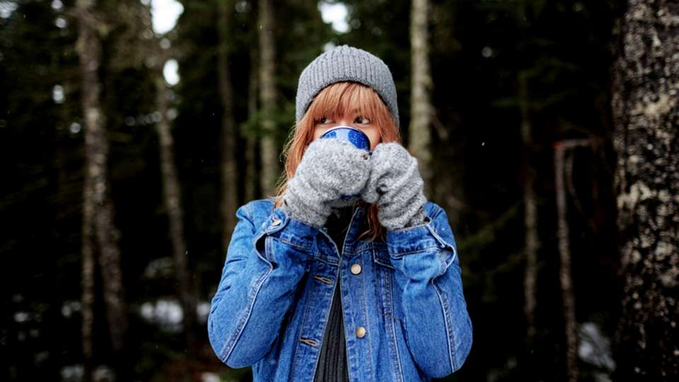 A woman wearing cold weather clothes sips a hot drink standing out in the woods.