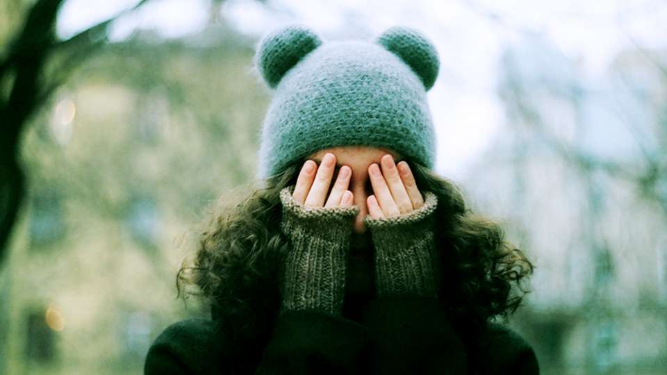 A person with long hair wearing a winter hat covers her face with her hands.