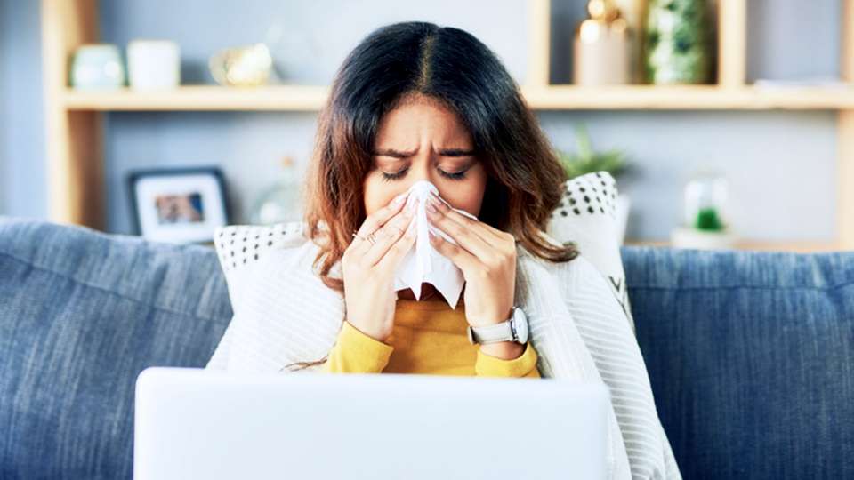 A woman sitting on her couch wrapped in a blanket blows her nose.