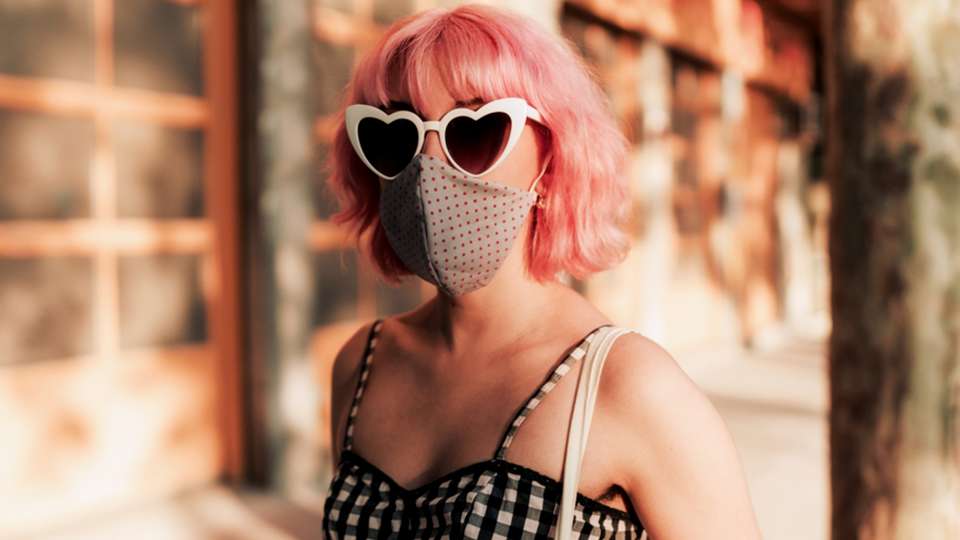 A light-skinned woman with short pink hair wears a face mask and heart-shaped sunglasses.