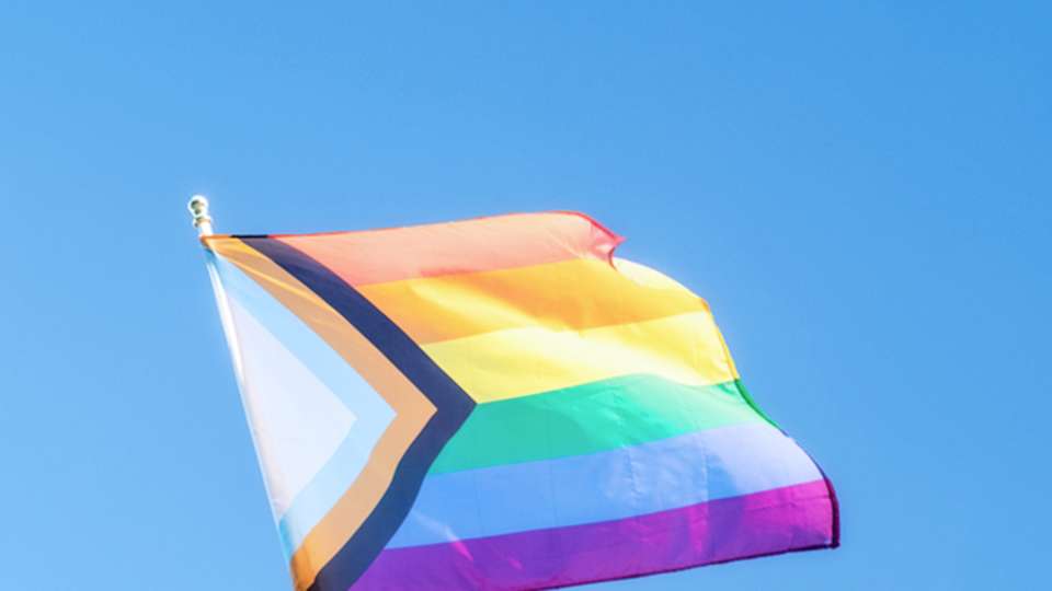 A progress pride flag flies against a blue sky.