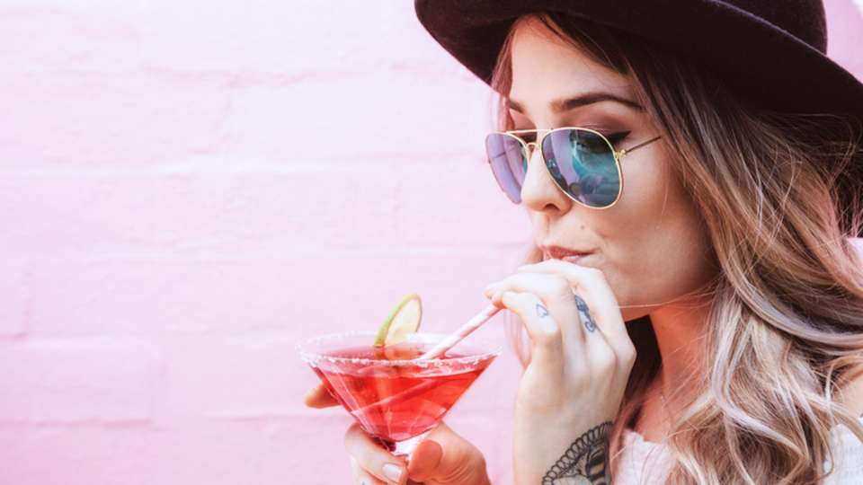 A white woman wearing a hat and sunglasses sips from a cocktail standing in front of a pink-painted brick wall.