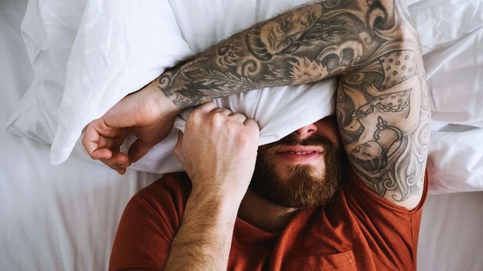 A man lies in bed with a pillow covering his face.