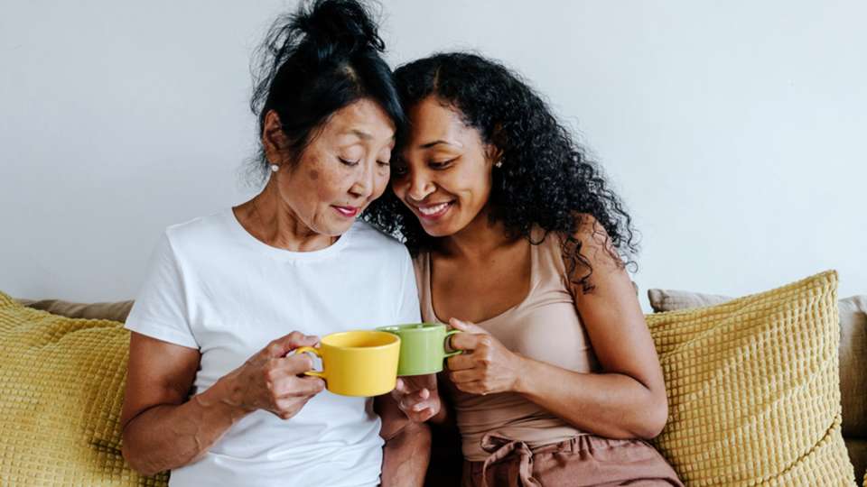 A mom and her adult daughter hold mugs while sitting on a couch.