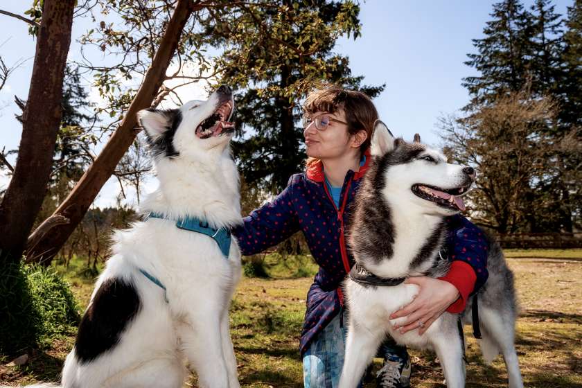 A woman kneeling outdoors with two husky-like dogs.