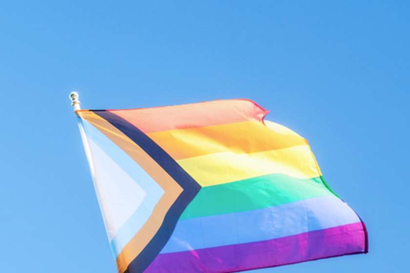 A progress pride flag flies against a blue sky.
