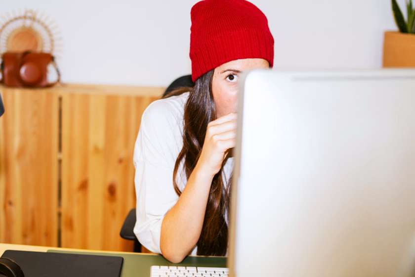 A woman in a red beanie peeks around a computer.