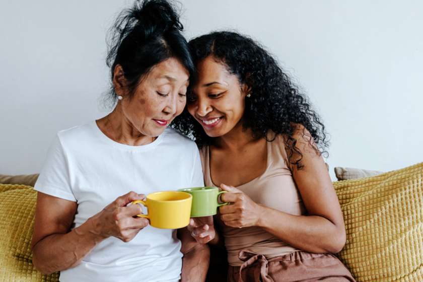 A mom and her adult daughter hold mugs while sitting on a couch.