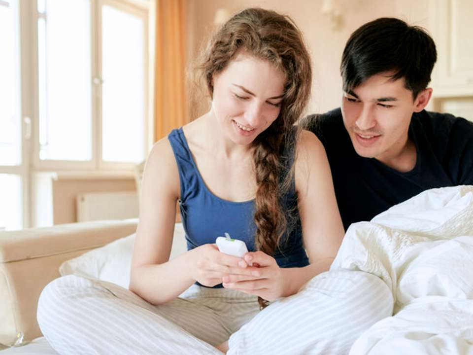 A man and woman sit in bed looking at the woman's glucose monitor.