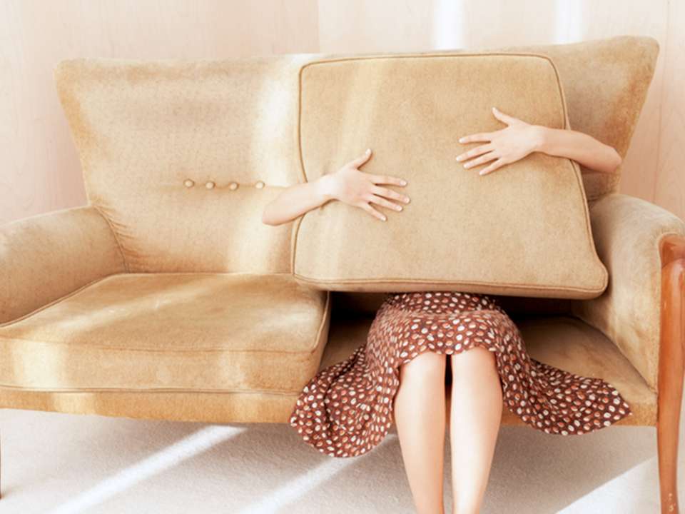 A woman sits on a couch holding a pillow over her upper body and face.