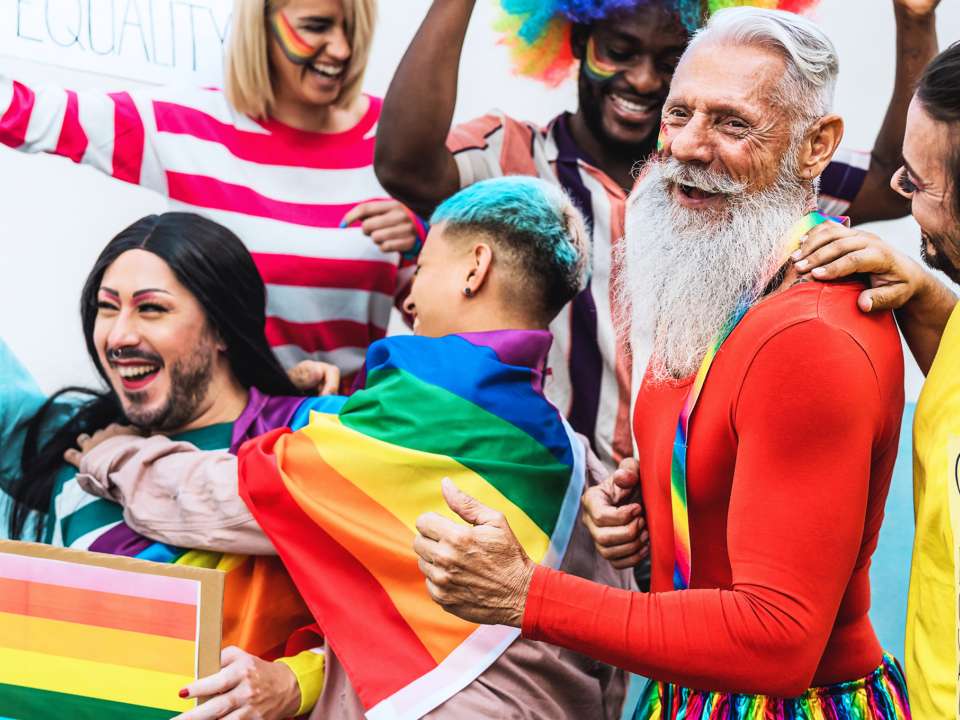 A smiling older adult is surrounded by people of all ages at a Pride event.