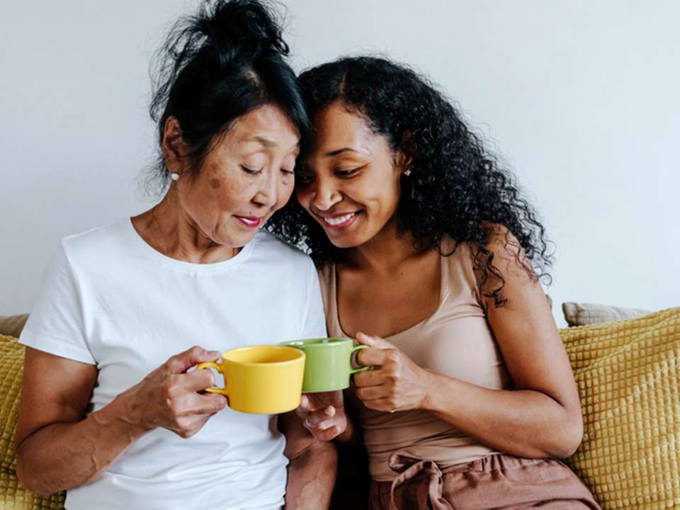 A mom and her adult daughter hold mugs while sitting on a couch.